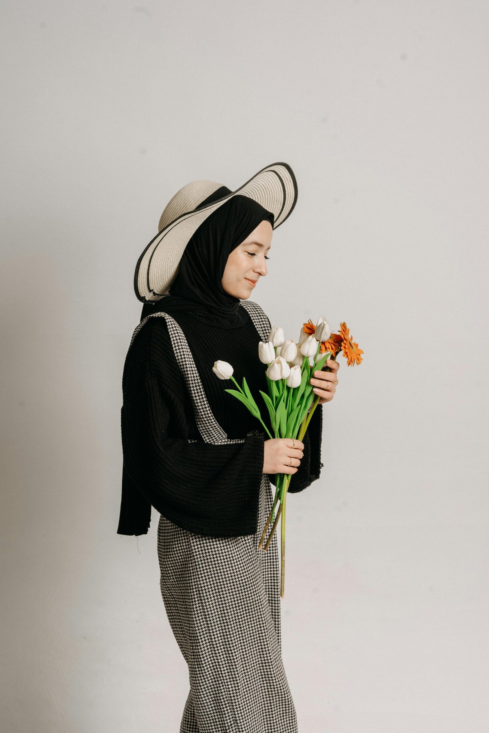 A stylish young woman wearing a hijab and hat holds white tulips and a daisy, posing indoors against a neutral background.